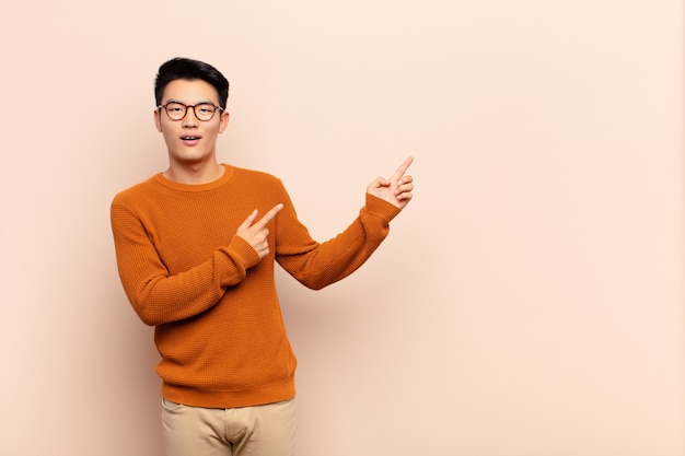 Joven chino que se siente alegre y sorprendido, sonriendo con una expresión de asombro y apuntando hacia un lado en la pared de color plano