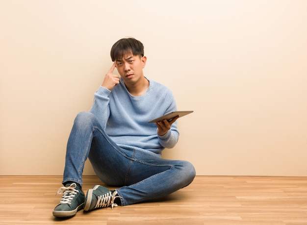 Joven chino sentado usando su tableta haciendo un gesto de concentración