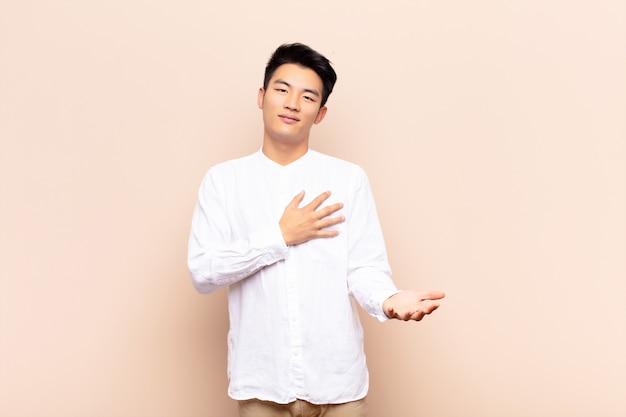 Joven chino sintiéndose feliz y enamorado, sonriendo con una mano al lado del corazón y la otra estirada al frente contra una pared de color plano