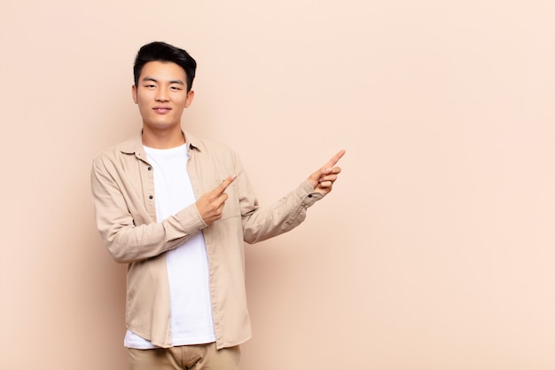 Joven chino sonriendo alegremente y apuntando hacia un lado y hacia arriba con ambas manos mostrando el objeto en el espacio de la copia contra la pared de color plano