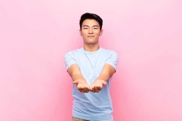 Joven chino sonriendo felizmente con mirada amigable, segura y positiva, ofreciendo y mostrando un objeto o concepto contra la pared de color