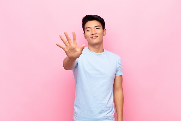 Joven chino sonriendo y mirando amigablemente, mostrando el número cinco o quinto con la mano hacia adelante, contando hacia atrás en la pared de color plano