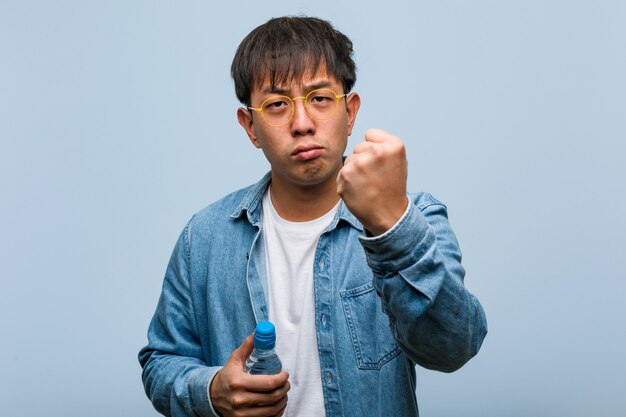 Joven chino sosteniendo una botella de agua mostrando el puño al frente, expresión enojada