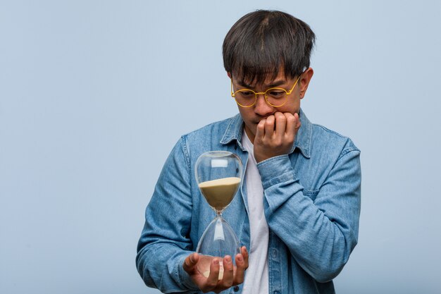 Foto joven chino sosteniendo un reloj de arena mordiéndose las uñas, nervioso y muy ansioso