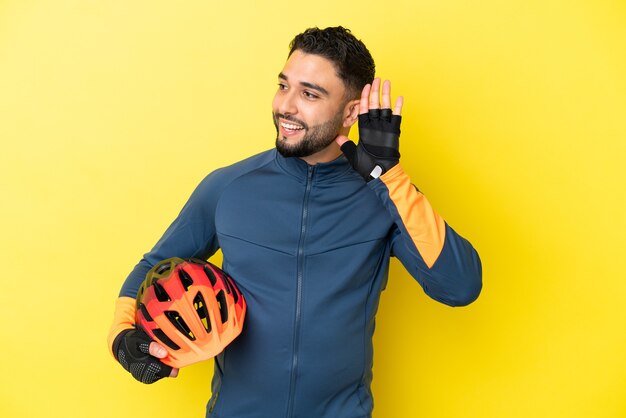 Joven ciclista árabe aislado sobre fondo amarillo escuchando algo poniendo la mano en la oreja