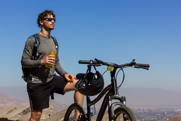 Joven ciclista con gafas de sol sosteniendo una botella de agua junto a la bicicleta de montaña en un día soleado