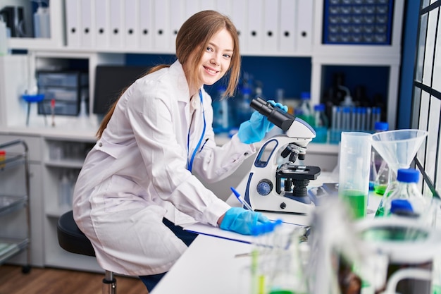 Joven científica caucásica que usa microscopio escribe en un documento en el laboratorio