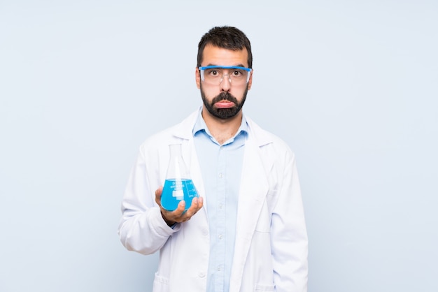 Joven científico sosteniendo frasco de laboratorio sobre pared aislada con expresión triste y deprimida