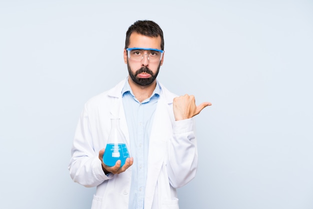 Joven científico sosteniendo frasco de laboratorio sobre pared aislada infeliz y apuntando hacia un lado