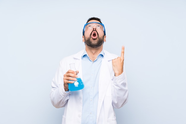 Joven científico sosteniendo frasco de laboratorio sobre pared aislada sorprendido y apuntando hacia arriba