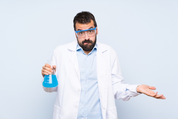 Joven científico sosteniendo frasco de laboratorio sobre pared aislada teniendo dudas al levantar las manos