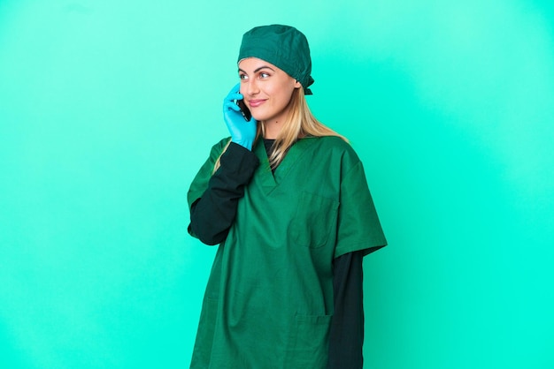 Joven cirujana uruguaya con uniforme verde aislada de fondo azul manteniendo una conversación con alguien por teléfono móvil