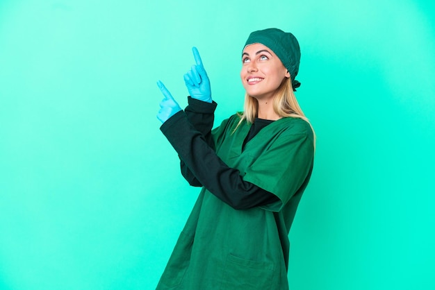 Joven cirujana uruguaya con uniforme verde aislada de fondo azul señalando con el dedo índice una gran idea