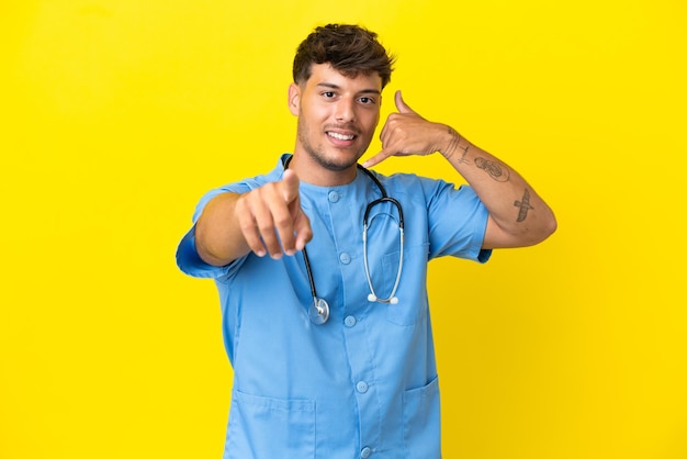 Joven cirujano médico hombre aislado sobre fondo amarillo haciendo gesto de teléfono y apuntando al frente