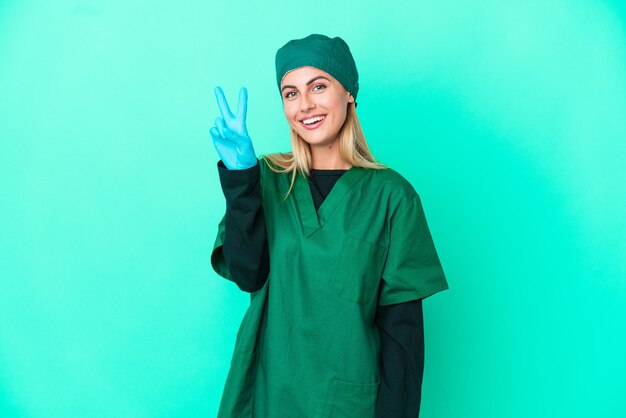 Joven cirujano mujer uruguaya en uniforme verde aislado sobre fondo azul sonriendo y mostrando el signo de la victoria