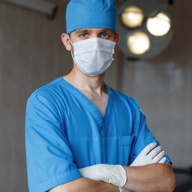 Joven cirujano en uniforme azul con una máscara blanca médica con guantes de goma está de pie en la sala de operaciones