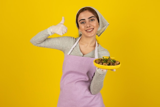 Joven cocinera en delantal sosteniendo un plato de champiñones fritos y mostrando los pulgares para arriba.