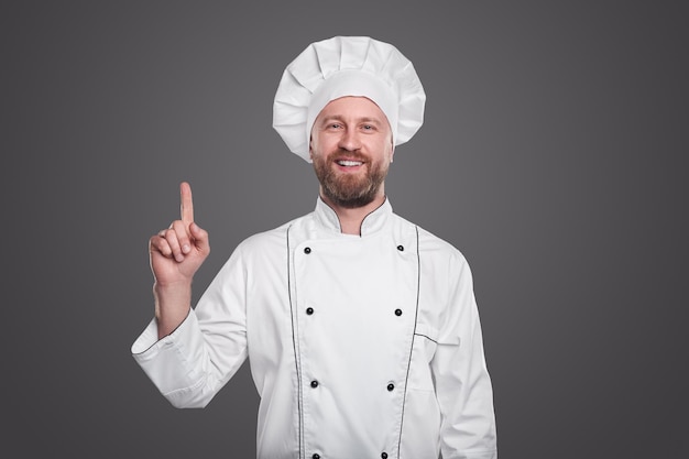 Joven cocinero en uniforme de chef blanco sonriendo y de pie sobre fondo gris mientras demuestra el pulgar hacia arriba gesto
