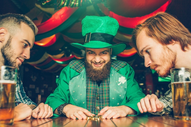 El joven codicioso en traje verde se sienta a la mesa con amigos y mira las monedas de oro que agarró. Otros chicos los miran también. Tazas de cerveza de pie sobre la mesa.