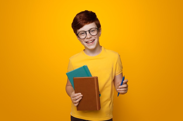 Joven colegial de jengibre sosteniendo algunas carpetas está sonriendo en una pared de estudio amarillo mientras usa gafas y camiseta casual