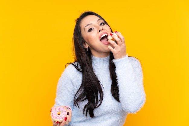 Joven colombiana sobre pared amarilla aislada comiendo una rosquilla