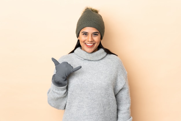 Joven colombiana con sombrero de invierno en pared beige haciendo gesto de teléfono