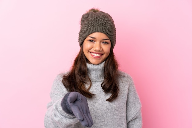 Joven colombiana con sombrero de invierno sobre pared rosa aislado apretón de manos después de un buen trato