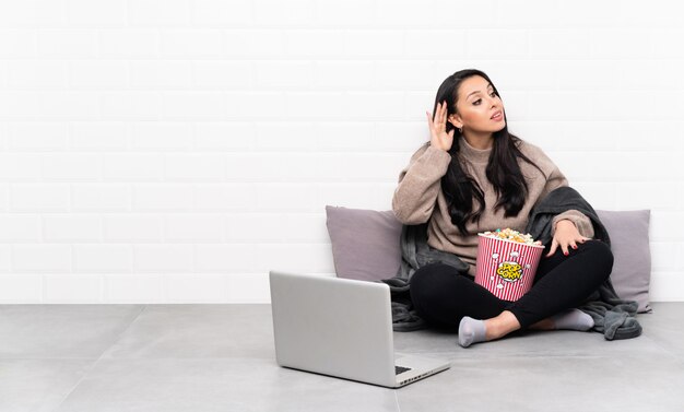 Joven colombiana sosteniendo un tazón de palomitas de maíz y mostrando una película en una computadora portátil escuchando algo poniendo la mano en la oreja