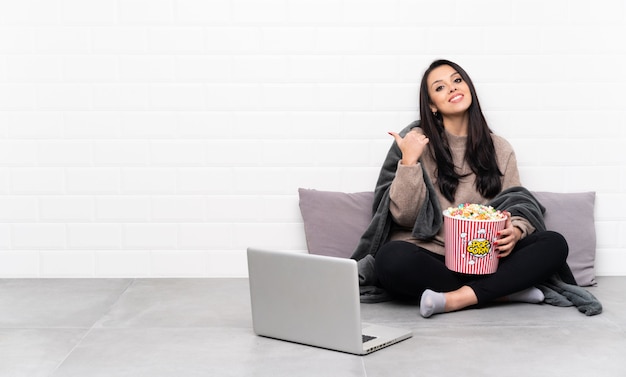 Joven colombiana sosteniendo un tazón de palomitas de maíz y mostrando una película en una computadora portátil con pulgares arriba gesto y sonriendo