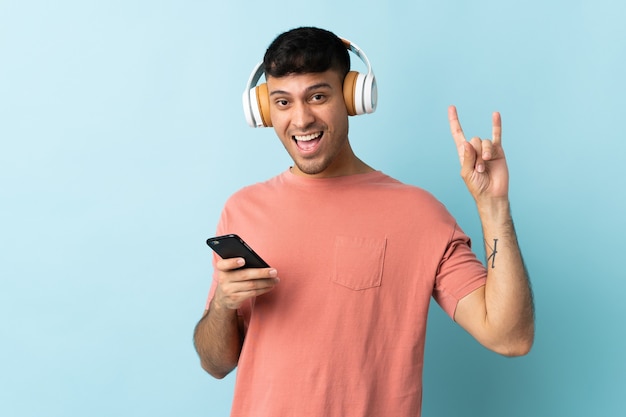 Joven colombiano aislado en azul escuchando música con un móvil haciendo gesto de rock