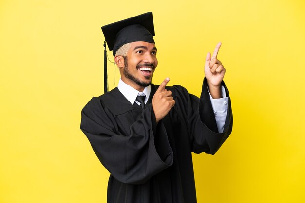 Joven colombiano graduado universitario aislado sobre fondo amarillo apuntando con el dedo índice una gran idea