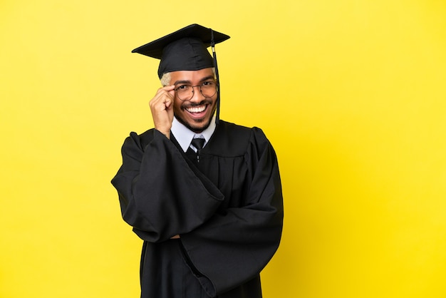 Joven colombiano graduado universitario aislado sobre fondo amarillo con gafas y feliz