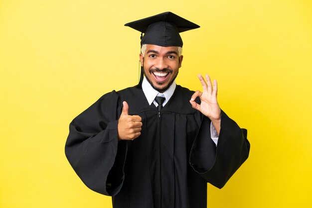 Joven colombiano graduado universitario aislado sobre fondo amarillo mostrando el signo de ok y el pulgar hacia arriba gesto