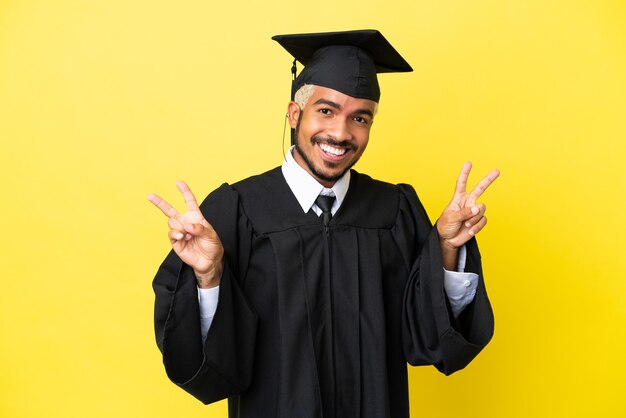 Joven colombiano graduado universitario aislado sobre fondo amarillo mostrando el signo de la victoria con ambas manos