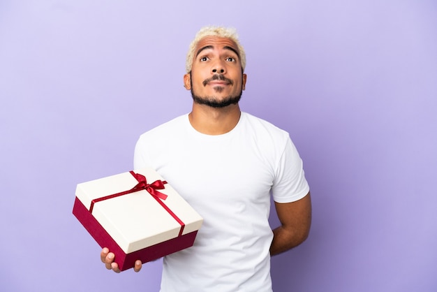 Joven colombiano sosteniendo un regalo aislado sobre fondo púrpura y mirando hacia arriba