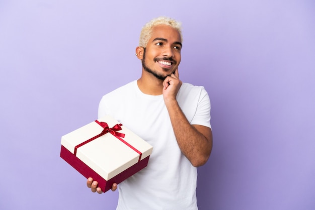 Joven colombiano sosteniendo un regalo aislado sobre fondo púrpura pensando en una idea mientras mira hacia arriba