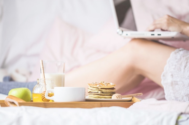 Joven come un desayuno saludable en la cama con un portátil