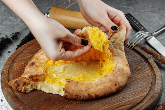 Una joven comienza a comer khachapuri fragante en ajarian con queso sobre un soporte de madera con una corteza crujiente.