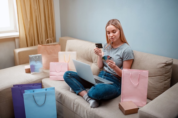 El joven comprador feliz con laptop está eligiendo la mejor tarjeta de crédito para el pago en la tienda en línea mientras está sentado en un sofá con bolsas de compras