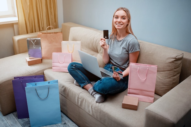 Joven comprador feliz con tarjetas de crédito está eligiendo la mejor venta mientras está sentado en un sofá con bolsas de compras