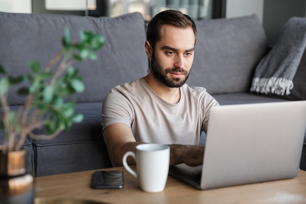 un joven concentrado en el interior de su casa usando una computadora portátil.