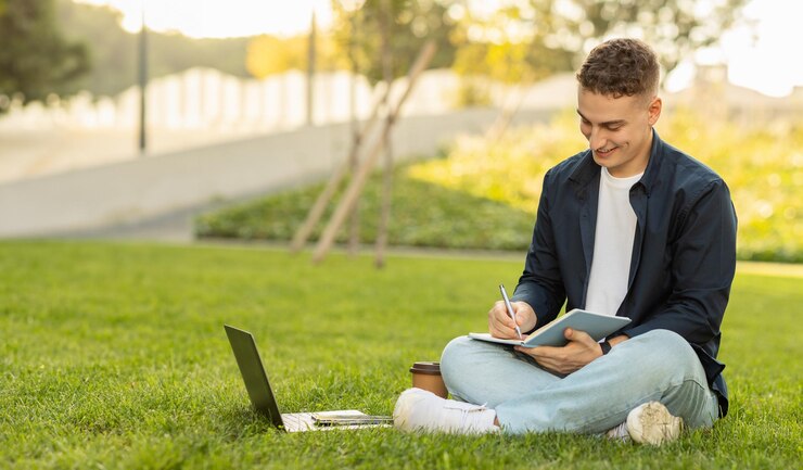 Un joven concentrado sentado en la hierba verde en un parque escribiendo en un cuaderno