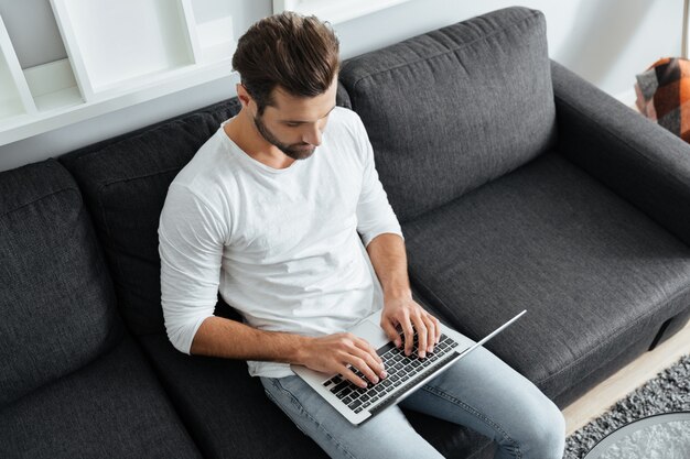 Joven concentrado sentado en el sofá usando la computadora portátil