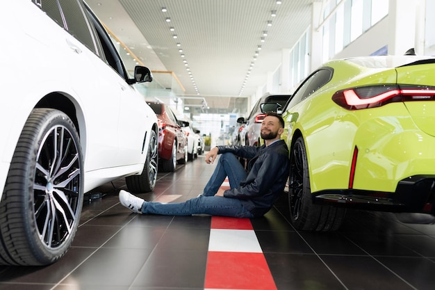 Un joven en un concesionario de automóviles se sienta en el suelo apoyado en un automóvil soñando con comprarlo