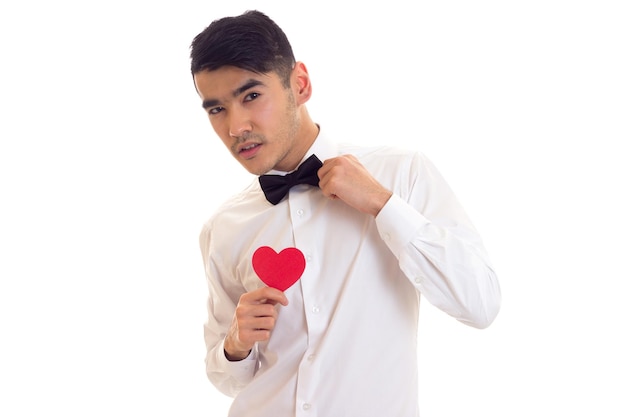 Joven confiado con cabello oscuro en camiseta blanca con pajarita negra sosteniendo un corazón de papel rojo sobre fondo blanco en el estudio