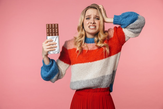 Joven confundido nervioso muy hermosa mujer posando aislado sobre pared rosa sosteniendo chocolate