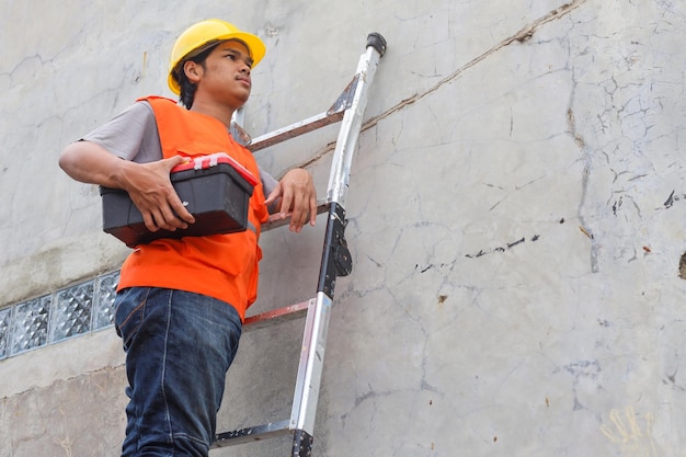 Joven constructor masculino con casco de seguridad amarillo y chaleco naranja mirando enfocado subiendo la escalera