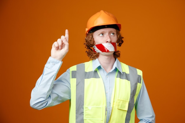 Joven constructor en uniforme de construcción y casco de seguridad con cinta adhesiva en la boca mirando hacia arriba mostrando el dedo índice de pie sobre fondo naranja