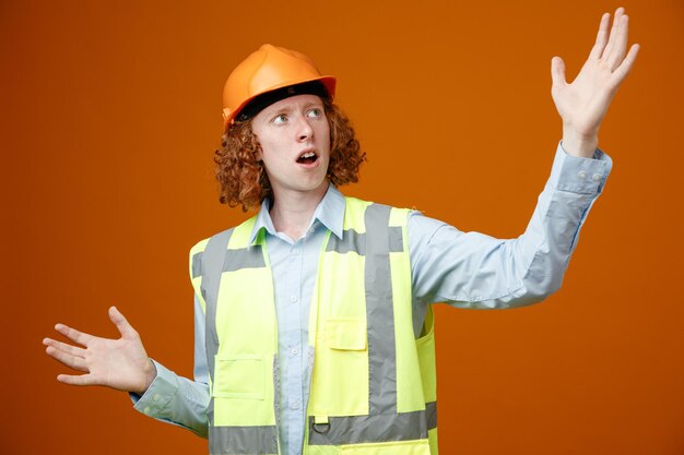 Joven constructor en uniforme de construcción y casco de seguridad mirando hacia arriba emocionado y gesticulando emocionalmente con las manos de pie sobre fondo naranja