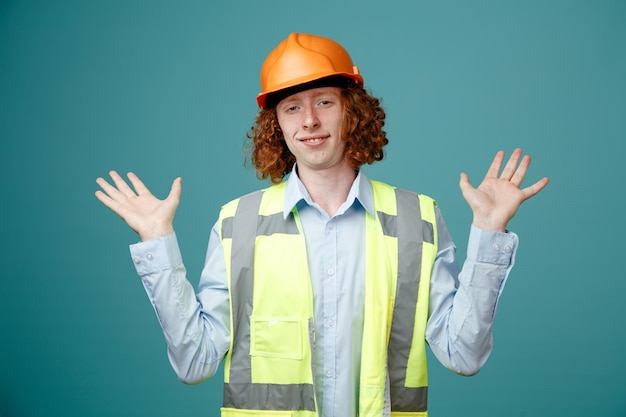 Foto joven constructor en uniforme de construcción y casco de seguridad mirando a la cámara confundido levantando los brazos sin respuesta de pie sobre fondo azul.